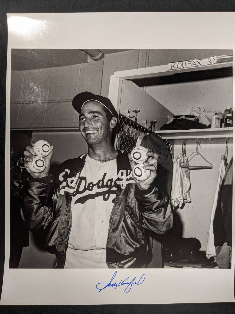 Black-and-white photo of Sandy Koufax smiling in a Dodgers locker room, holding four baseballs marked with zeros for his four no-hitters, next to a locker labeled Koufax 32